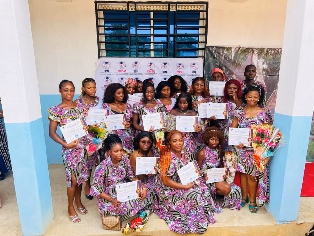 La promotion au complet, en tenue de pagne assortie, pose sous le préau devant le kakémono IFPPB et EFOPSAB, attestations et bouquets à la main.