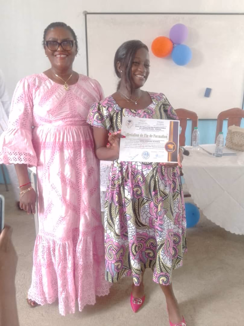 Une lauréate présente son attestation de fin de formation aux côtés d'une responsable de l'institut, devant le tableau décoré de ballons.