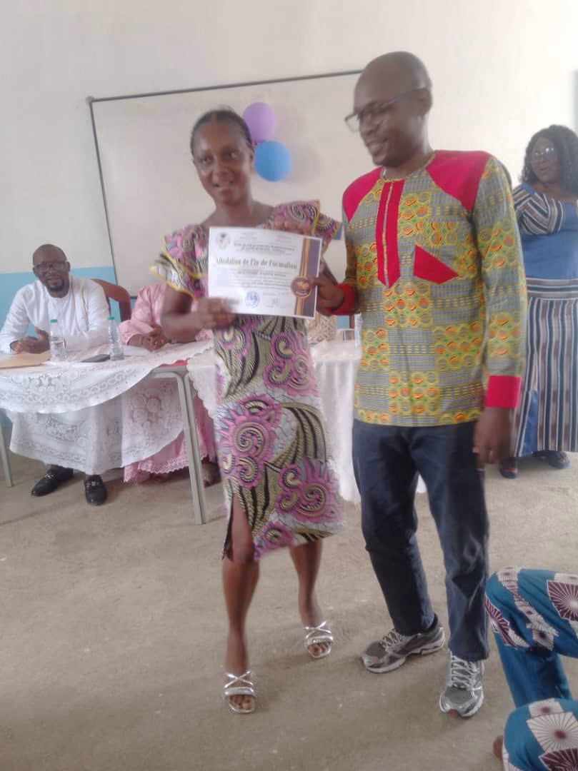 Une lauréate tient son attestation ouverte à deux mains, accompagnée d'un membre de l'encadrement en tenue traditionnelle.