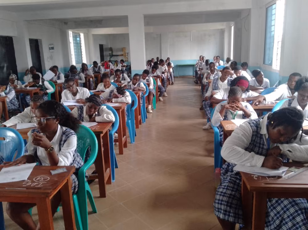 Une salle de classe carrelée où les apprenantes en uniforme composent, installées à des tables individuelles en bois.